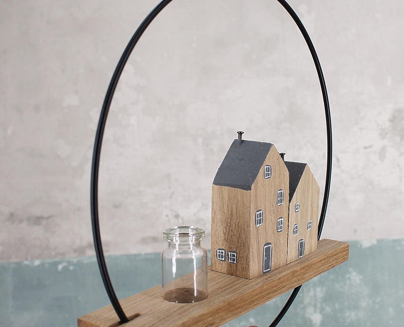 May include: A decorative wall hanging featuring a thin black metal hoop attached to a horizontal wooden plank. On the plank sits a small clear glass bottle and two miniature wooden house figures with dark gray roofs and painted white windows.