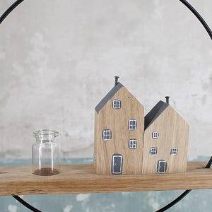 May include: A decorative wall hanging featuring a thin black metal circle frame holding a horizontal wooden shelf. On the shelf sits a small clear glass bottle and two wooden house-shaped blocks with painted gray roofs, windows, and doors.