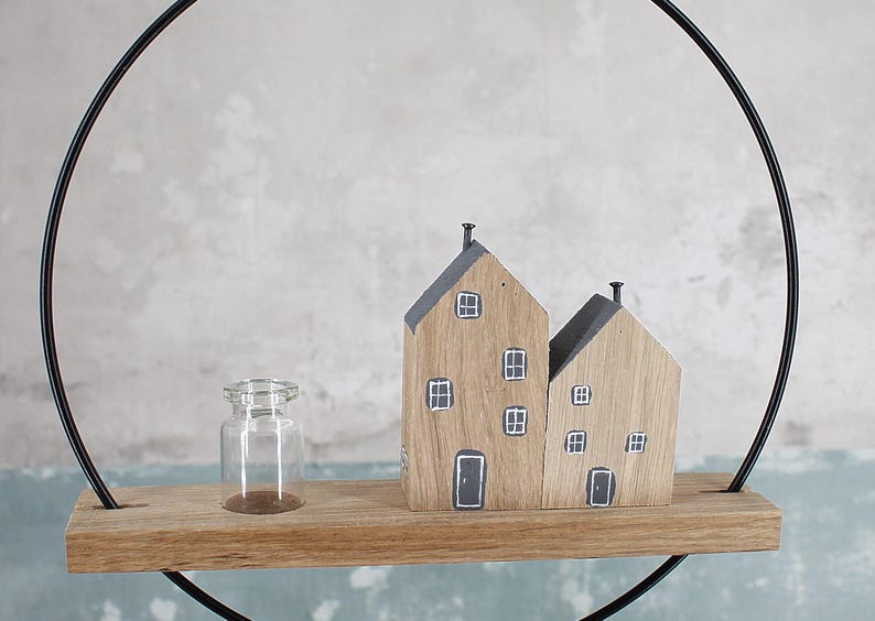 May include: A decorative wall hanging featuring a thin black metal circle frame holding a horizontal wooden shelf. On the shelf sits a small clear glass bottle and two wooden house-shaped blocks with painted gray roofs, windows, and doors.