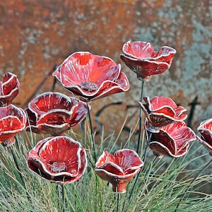 May include: A collection of ten ceramic poppy flowers with glossy red petals and white-trimmed edges, each mounted on a thin black metal stem. The flowers are arranged in a cluster, showcasing varying sizes and open, bowl-shaped blooms.