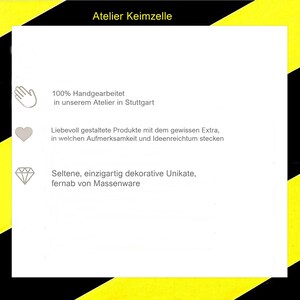 May include: A white square sign with a yellow and black diagonal striped border. The text reads Atelier Keimzelle, followed by three lines of German text accompanied by icons of a hand, a heart, and a diamond, detailing production.