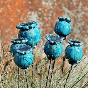 May include: A collection of six ceramic poppy seed pods in a vibrant turquoise blue glaze with a distinct crackle finish. Each rounded pod features a scalloped top and is mounted on a thin, dark metal stake, creating a sculptural garden display.