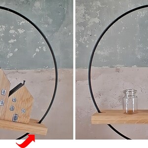 May include: A circular black metal frame holds a horizontal wooden shelf. On the shelf sit two wooden house-shaped blocks with painted windows and doors, alongside a small, clear glass jar. The shelf is suspended within the metal ring.