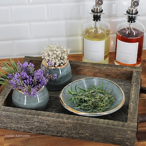 May include: A dark brown wooden tray containing two small blue-grey ceramic pots with gold rims, one filled with lavender and one with baby's breath, plus a matching shallow bowl holding fresh herbs, near two glass oil and vinegar bottles.