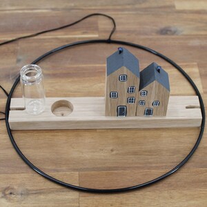 May include: A decorative wall hanging featuring a black metal hoop, a light wood rectangular base, two small wooden house figurines with gray roofs and painted windows, and a clear glass vial attached to the wooden base.