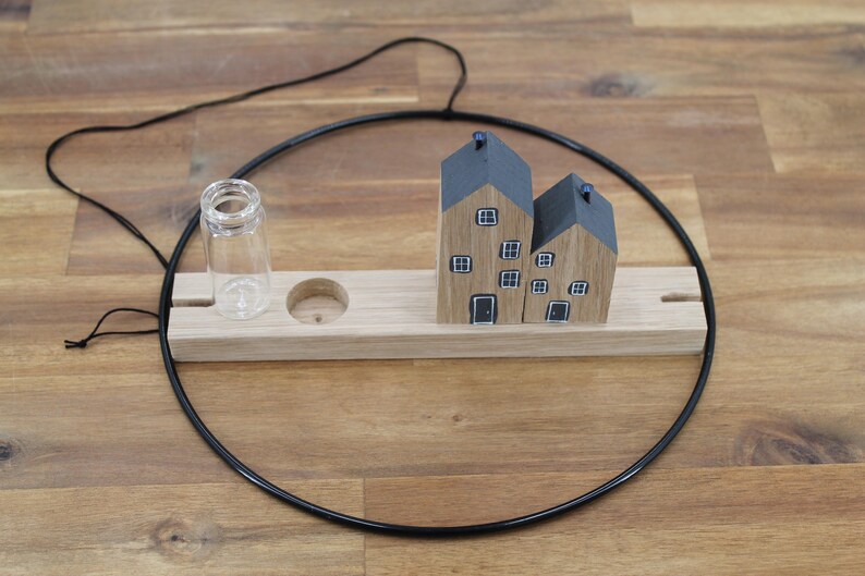 May include: A decorative wall hanging featuring a black metal hoop, a light wood rectangular base, two small wooden house figurines with gray roofs and painted windows, and a clear glass vial attached to the wooden base.