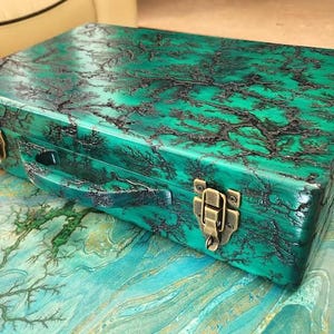 May include: A rectangular, emerald green box with a black, tree-like pattern. The box has a handle and brass-colored clasps. The box is sitting on a teal and blue surface.