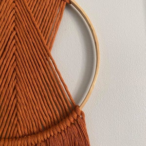 May include: A macrame wall hanging with a geometric pattern in a burnt orange colour. The hanging is made with thick cotton cord and has a wooden hoop at the top.