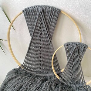May include: Two grey macrame wall hangings with fringe, each hanging from a wooden hoop. The larger hoop is approximately 30 cm in diameter, and the smaller hoop is approximately 20 cm in diameter.