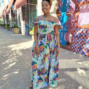 Puede incluir: Un vestido maxi blanco con estampado floral colorido. El vestido tiene un escote de hombros descubiertos con volantes y un corpi&ntilde;o ajustado. La falda es amplia y fluida.