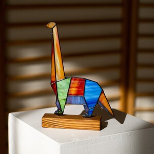 May include: A colorful stained glass dinosaur sculpture with a long neck and tail. The dinosaur is made of blue, green, red, orange, and purple glass. It is standing on a wooden base.