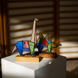 May include: Two colorful stained glass dinosaur figurines, one a long-necked dinosaur and the other a three-horned dinosaur, are displayed on a white pedestal.