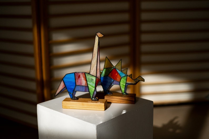 May include: Two colorful stained glass dinosaur figurines, one a long-necked dinosaur and the other a three-horned dinosaur, are displayed on a white pedestal.