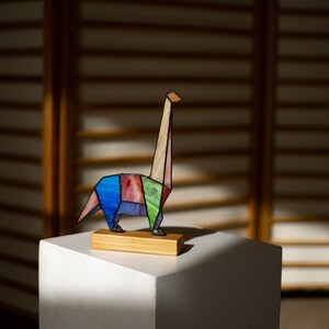 May include: A colorful stained glass dinosaur sculpture with a long neck, standing on a wooden base. The dinosaur is made of various shades of blue, green, pink, and brown glass.