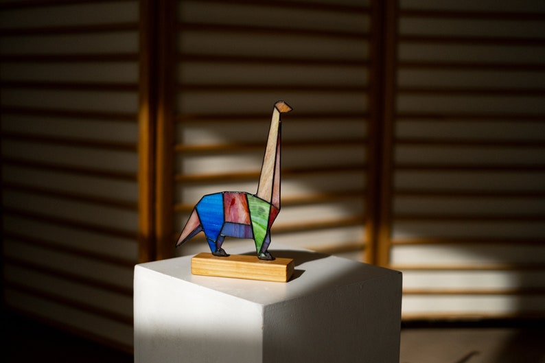 May include: A colorful stained glass dinosaur sculpture with a long neck, standing on a wooden base. The dinosaur is made of various shades of blue, green, pink, and brown glass.