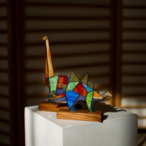 May include: A colorful stained glass dinosaur sculpture with a long neck and spikes on its back. The sculpture is displayed on a wooden base and a white pedestal.