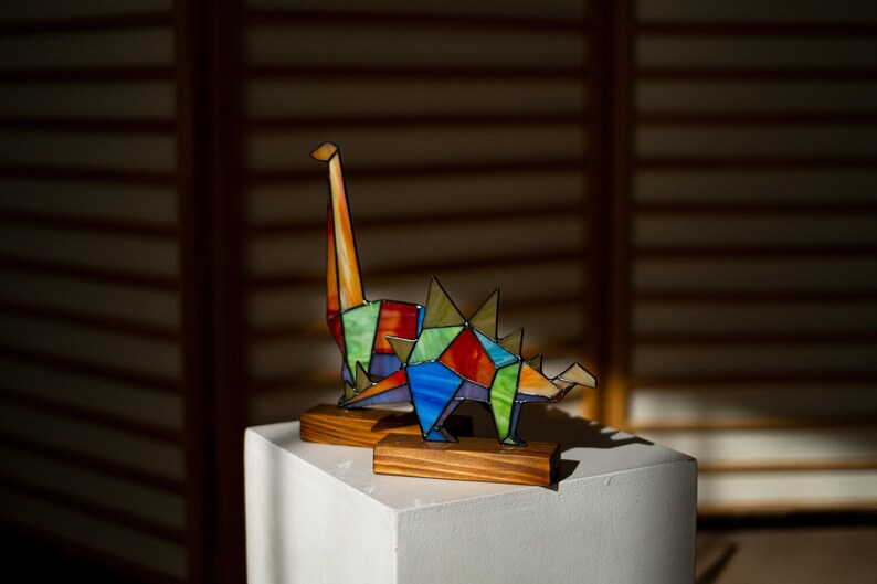 May include: A colorful stained glass dinosaur sculpture with a long neck and spikes on its back. The sculpture is displayed on a wooden base and a white pedestal.
