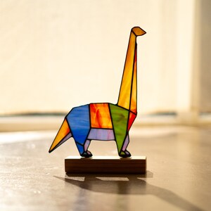 May include: A colorful stained glass origami dinosaur sculpture on a wooden base. The dinosaur is yellow, orange, green, blue, and purple.