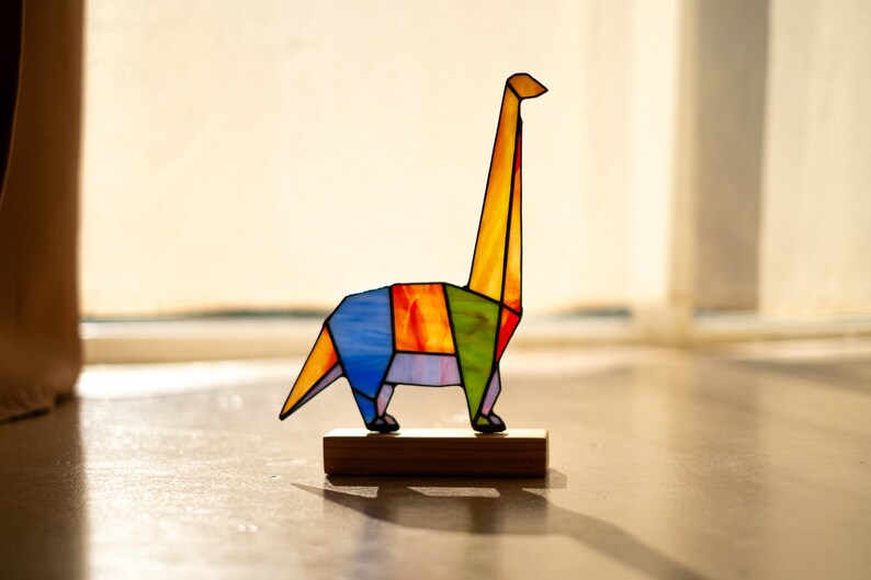 May include: A colorful stained glass origami dinosaur sculpture on a wooden base. The dinosaur is yellow, orange, green, blue, and purple.