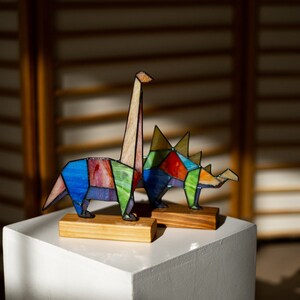May include: Two colorful stained glass dinosaur figurines, one a long-necked dinosaur and the other a three-horned dinosaur, are displayed on a white pedestal.