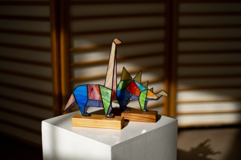 May include: Two colorful stained glass dinosaur figurines, one a long-necked dinosaur and the other a three-horned dinosaur, are displayed on a white pedestal.