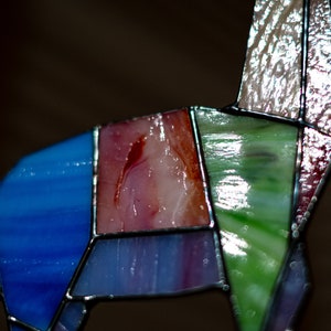 May include: A stained glass sculpture of a stylized animal, possibly a deer or a horse. The sculpture is made of various colors of glass, including blue, green, pink, and red. The glass pieces are joined together with black metal.