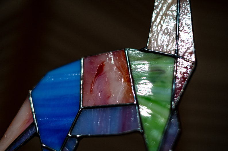 May include: A stained glass sculpture of a stylized animal, possibly a deer or a horse. The sculpture is made of various colors of glass, including blue, green, pink, and red. The glass pieces are joined together with black metal.