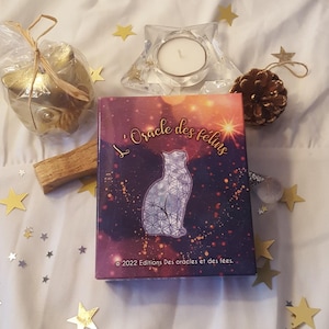 Op de afbeelding: Een rechthoekige orakelkaarten doos getiteld L'Oracle des félins met een kat-silhouet met een geometrisch patroon tegen een paarse en oranje nevelachtergrond, met de tekst 2022 Editions Des oracles et des fées onderaan gedrukt.