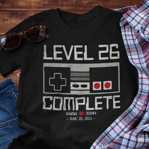 May include: Black t-shirt with a white graphic of a video game controller and the text "Level 26 Complete". The graphic also includes a heart and the text "Sara John - June 20, 2021 - "