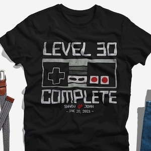 May include: Black t-shirt with a retro video game controller graphic and the text "Level 30 Complete" with a heart and the names "Sady & John" and the date "June 20, 2021".