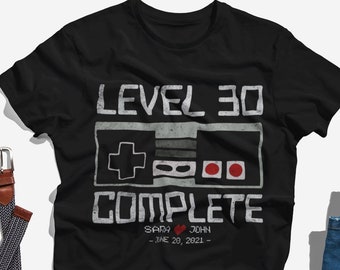 Op de afbeelding: Zwart T-shirt met een retro videogamecontroller-afbeelding en de tekst "Level 30 Complete" met een hartje en de namen "Sady & John" en de datum "20 juni 2021".
