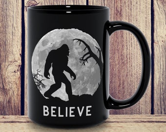 Può includere: Tazza da caffè nera con una silhouette bianca di Bigfoot che cammina davanti a una luna piena. Il testo "BELIEVE" è stampato sotto la luna.