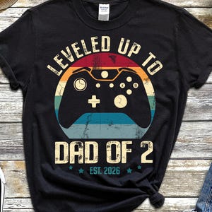 Puede incluir: Camiseta negra con un gráfico colorido de un mando de videojuegos y el texto "LEVELED UP TO DAD OF 2 EST. 2026". La camiseta se combina con pantalones cortos vaqueros azules y zapatillas negras.