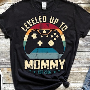 May include: Black t-shirt with a retro-style video game controller graphic. The design includes the text "LEVELED UP TO MOMMY EST. 2026". The shirt is paired with denim shorts and black sneakers.