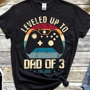 Könnte beinhalten: Schwarzes T-Shirt mit einem Retro-Videospiel-Controller-Motiv. Der Text lautet "LEVELED UP TO DAD OF 3 EST. 2026". Das Shirt wird mit blauen Jeansshorts und schwarzen Turnschuhen kombiniert. Ein Strohhut und eine Uhr sind ebenfalls zu sehen.