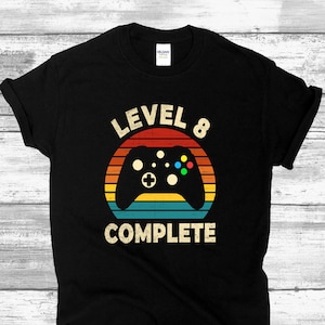May include: Black t-shirt with a retro-style graphic of a video game controller in a rainbow circle. The text "LEVEL 8 COMPLETE" is below the circle.