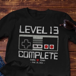 May include: Black t-shirt with a white graphic of a video game controller and the text "Level 13 Complete" with the names "Sara & John" and the date "June 28, 2021".