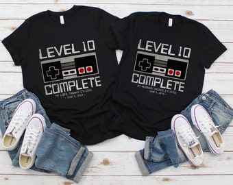 May include: Two black t-shirts with a graphic of a video game controller and the text "Level 10 Complete". The left shirt says "My wife thinks I'm cool - June 5, 2013 -" and the right shirt says "My husband thinks I'm cute - June 5, 2013 -"