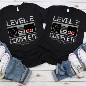 May include: Two black t-shirts with a video game controller graphic and the text "Level 2 Complete". The left shirt says "My wife thinks I'm cool!" and the right shirt says "My husband thinks I'm cute!" Both shirts have the date "June 2, 2021".