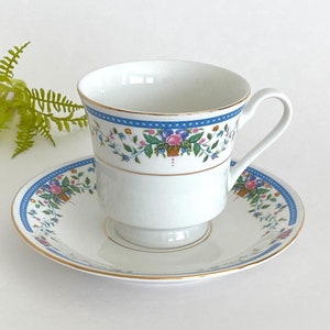 Könnte beinhalten: Ein weißes Porzellan-Teetassen- und Untertassen-Set mit einem Blumenmuster in Blau-, Rosa- und Grüntönen. Die Tasse hat einen goldenen Rand und die Untertasse hat einen goldenen Streifen am Rand.