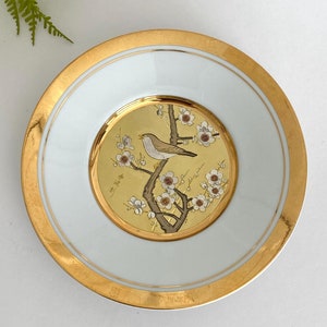 May include: A white porcelain plate with a gold rim and a gold-plated image of a bird perched on a branch with white blossoms. The plate is a decorative piece with a Japanese theme.