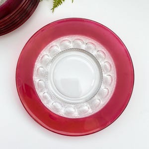 May include: A stack of red-rimmed glass plates with a clear, textured center and a decorative border. The plates are arranged on a white surface, with a sprig of green foliage in the background. The plates are a decorative tableware item.
