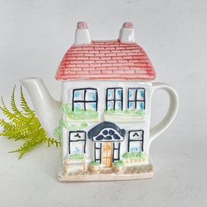 Puede incluir: Tetera de cerámica blanca con forma de casa con techo rojo, jardineras verdes y una puerta marrón. La tetera tiene un pico y una asa.