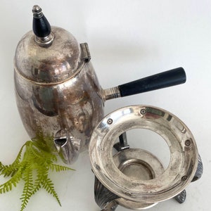 Puede incluir: Cafetera de plata con mango negro y soporte de plata. La cafetera tiene un pomo decorativo en la tapa.