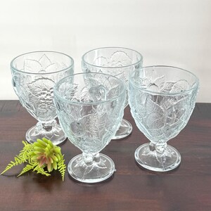 May include: Four clear glass goblets with a detailed embossed leaf and fruit design. The goblets have a stemmed base and a wide bowl. A small green and pink floral accent is in the foreground. The goblets are arranged on a dark brown surface.