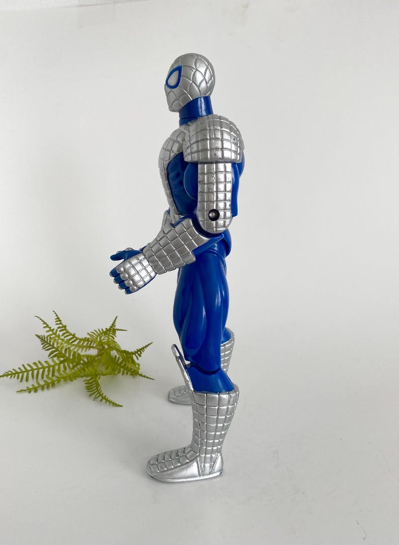 K&ouml;nnte beinhalten: Eine blaue und silberne Actionfigur von Spider-Man, die eine silberne R&uuml;stung tr&auml;gt. Die Figur ist mit erhobenem rechten Arm und dem linken Arm, der im Ellbogen gebeugt ist, aufgestellt.