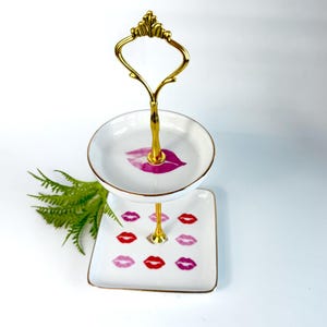 May include: Two-tiered serving tray with a gold-colored handle and support. The top tier is round, and the bottom tier is square. Both tiers are white with gold trim and feature lip print designs in red and pink.