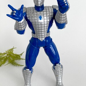 K&ouml;nnte beinhalten: Eine blaue und silberne Actionfigur von Spider-Man, die eine silberne R&uuml;stung tr&auml;gt. Die Figur hat ein Spinnenlogo auf der Brust und ist mit ausgestreckten Armen posiert.