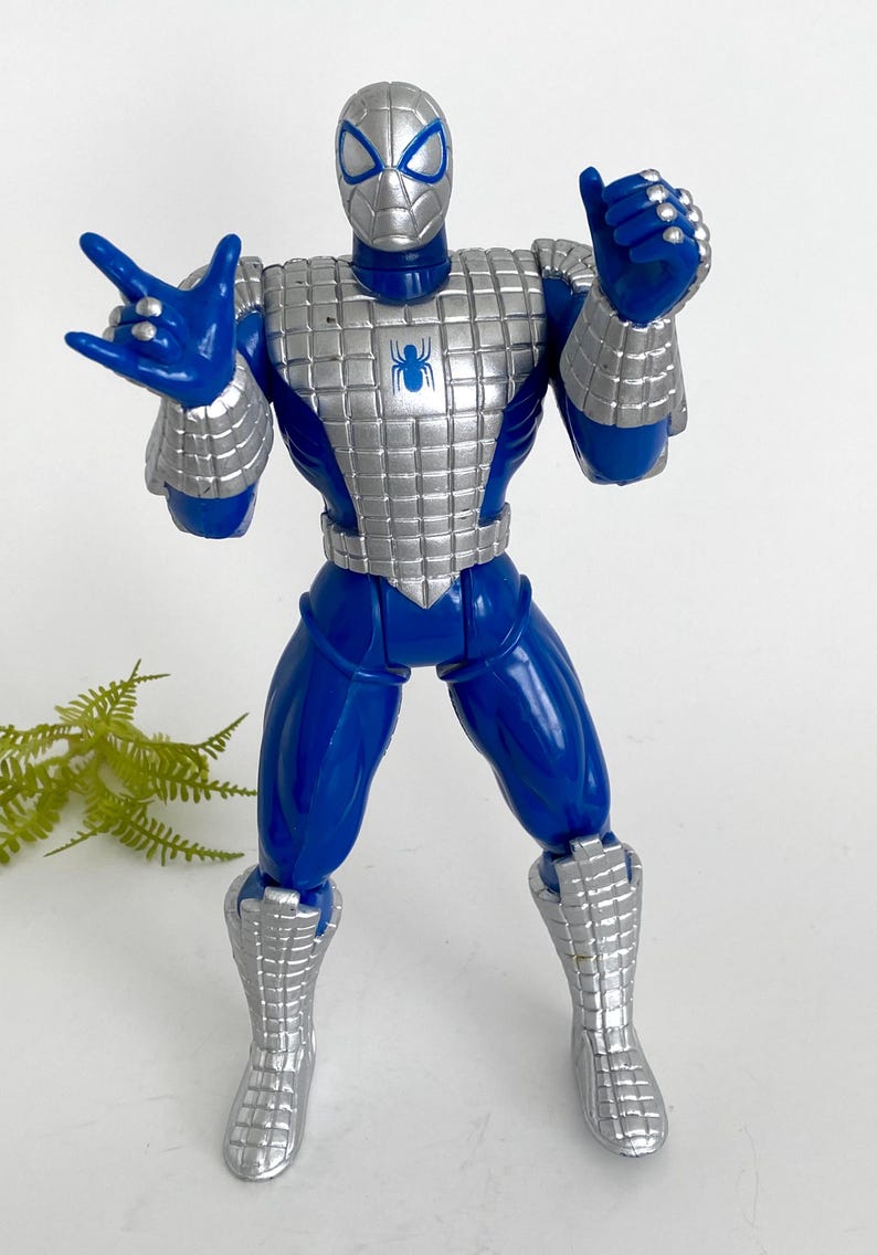 K&ouml;nnte beinhalten: Eine blaue und silberne Actionfigur von Spider-Man, die eine silberne R&uuml;stung tr&auml;gt. Die Figur hat ein Spinnenlogo auf der Brust und ist mit ausgestreckten Armen posiert.