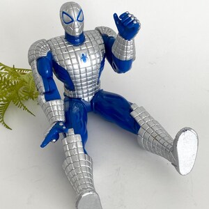 K&ouml;nnte beinhalten: Eine blaue und silberne Actionfigur von Spider-Man, die eine silberne R&uuml;stung tr&auml;gt. Die Figur sitzt mit dem rechten Arm erhoben in einer Daumen-hoch-Geste.